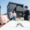 新しい積水ハウスの前で、不動産担当者が若い日本人夫婦に家の説明をしている。手前には契約書や鍵、貯金箱が置かれている。