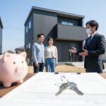 新しい積水ハウスの前で、不動産担当者が若い日本人夫婦に家の説明をしている。手前には契約書や鍵、貯金箱が置かれている。